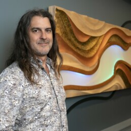 Featured artist Aaron Laux standing to the left of his artwork haning on a wall.