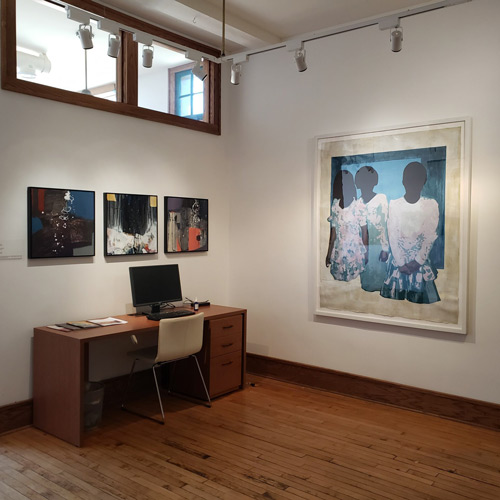 An art gallery with a desk and computer on the left wall with art hanging above it, and a large painting hanging on the adjacent wall